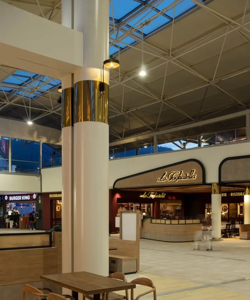 Interior de centro comercial moderno con zona de restauración, techos altos y estructura metálica vista