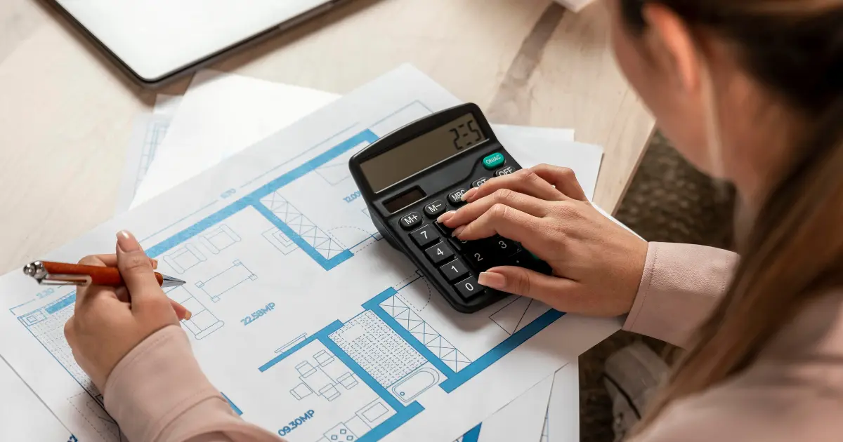person calculating budget on architectural plans with calculator on desk